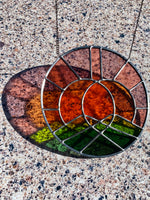 Stained glass sun catcher with brown, green, and orange colors on a speckled concrete surface.